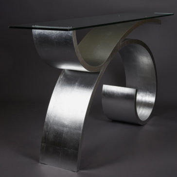 Exclusive Silver Leaf Console Table The Exclusive Silver Leaf Console Table boasts a modern design with an eye-catching contemporary glass surface. It features a curved, metallic base with a shimmering silver finish that loops at the bottom to support the sleek rectangular glass tabletop. The dark gradient background accentuates the table's striking form.
