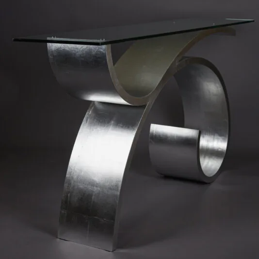Exclusive Silver Leaf Console Table The Exclusive Silver Leaf Console Table boasts a modern design with an eye-catching contemporary glass surface. It features a curved, metallic base with a shimmering silver finish that loops at the bottom to support the sleek rectangular glass tabletop. The dark gradient background accentuates the table's striking form.