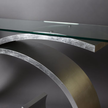Exclusive Silver Leaf Console Table Close-up of the Exclusive Silver Leaf Console Table, featuring a transparent glass top and a curved metal base. The design boasts sleek, clean lines and a modern aesthetic, set against a plain, dark background.