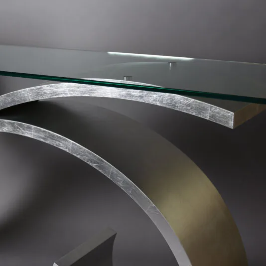 Exclusive Silver Leaf Console Table Close-up of the Exclusive Silver Leaf Console Table, featuring a transparent glass top and a curved metal base. The design boasts sleek, clean lines and a modern aesthetic, set against a plain, dark background.