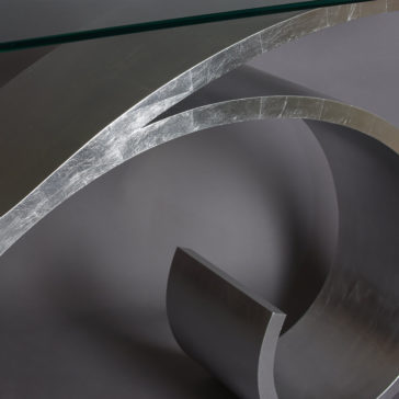 Exclusive Silver Leaf Console Table A close-up image of the Exclusive Silver Leaf Console Table features concentric metallic arcs with a sleek, reflective silver finish. The table is set against a neutral gray background, showcasing its glass surface atop the elegant arcs.