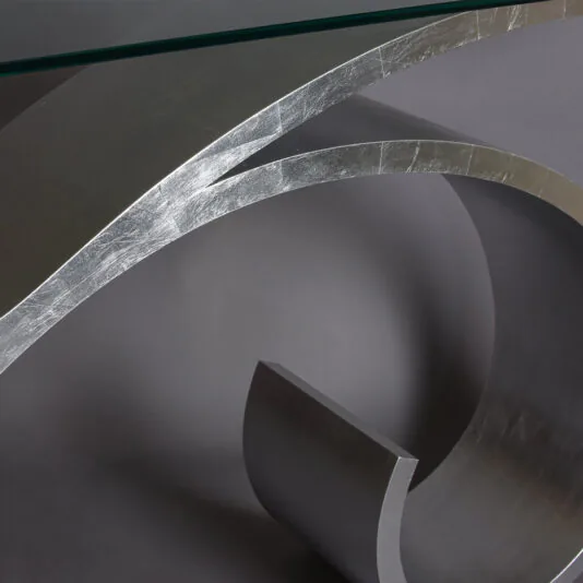 Exclusive Silver Leaf Console Table A close-up image of the Exclusive Silver Leaf Console Table features concentric metallic arcs with a sleek, reflective silver finish. The table is set against a neutral gray background, showcasing its glass surface atop the elegant arcs.