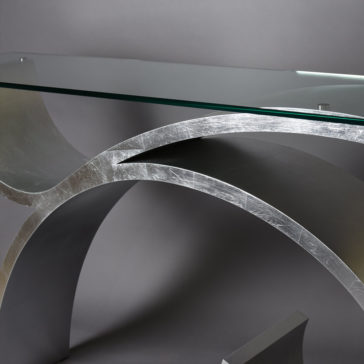 Exclusive Silver Leaf Console Table A close-up of the Exclusive Silver Leaf Console Table showcasing its sleek and artistic design. It features a clear glass top and a metallic, curved base with a brushed silver finish, set against a plain grey background.
