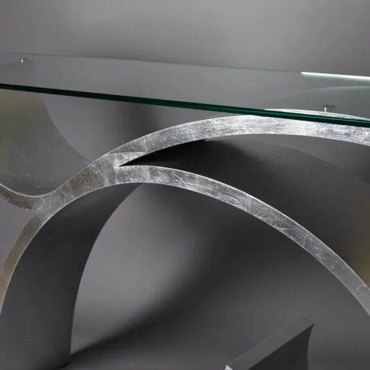 Exclusive Silver Leaf Console Table A close-up of the Exclusive Silver Leaf Console Table showcasing its sleek and artistic design. It features a clear glass top and a metallic, curved base with a brushed silver finish, set against a plain grey background.