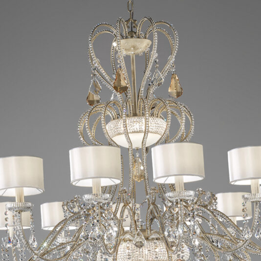 Large Crystal Chandelier With Shades The Large Crystal Chandelier With Shades is an ornate fixture that showcases multiple tiers of intricate embellishments and white cylindrical lampshades. It features a combination of curved metal arms and hanging crystal prisms, creating a striking illumination against a gray background.