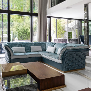 Leather Button Upholstered Corner Sofa A modern living room features a prominent Leather Button Upholstered Corner Sofa and matching cushions, complemented by a wooden coffee table with a marble top. Floor-to-ceiling windows allow for an expansive view of the lush greenery outside, while high ceilings and contemporary decor complete the sophisticated ambiance.