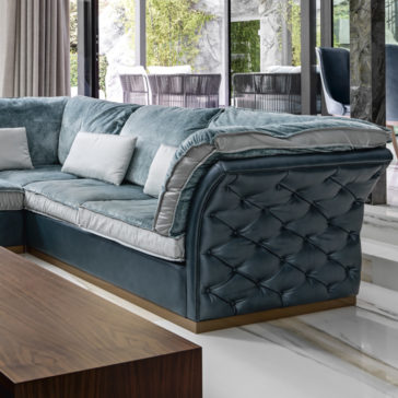 Leather Button Upholstered Corner Sofa A luxurious living room features the Leather Button Upholstered Corner Sofa, showcasing plush, deep teal leather with tufted accents and light grey cushioning. White throw pillows are arranged on the sofa. The room has large windows revealing a green garden patio with outdoor furniture. A sleek wooden coffee table is in the foreground.