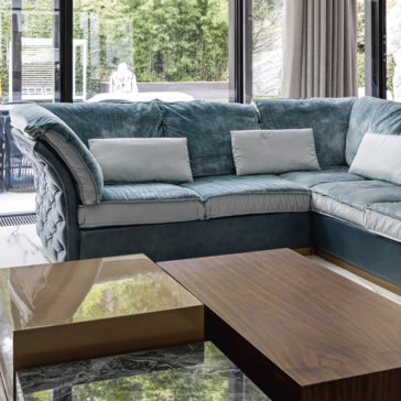 Leather Button Upholstered Corner Sofa A modern living room features a blue velvet sectional sofa adorned with white accent pillows. The Leather Button Upholstered Corner Sofa is positioned near expansive glass windows that reveal a view of lush greenery outside. In the foreground, two elegant coffee tables with marble and wood finishes are visible.