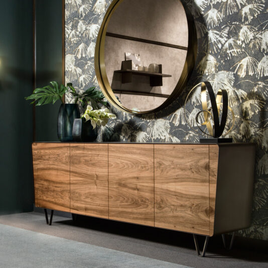 A modern living room with the Mid Century Style Walnut Sideboard featuring sleek, clean lines and metal legs. Above it, there's a large circular mirror reflecting the room. The sideboard is decorated with vases containing green leaves and white flowers, along with a sculpture to the right. A patterned wallpaper with leaf motifs adorns the background.