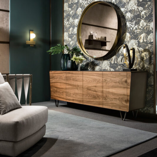 A modern living room showcasing the Mid Century Style Walnut Sideboard against a wallpapered wall adorned with leafy patterns. Above the sideboard, a large round mirror is perfectly centered, flanked by decorative vases and an abstract sculpture. In the foreground, there's a gray sofa complemented by a patterned wall light.