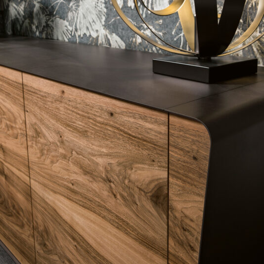 A close-up of the Mid Century Style Walnut Sideboard showcases its sleek, dark metal edges and natural wood grain doors. Atop the sideboard sits a contemporary sculpture featuring intertwined golden elements on a black base, all set against a wallpaper with a monochromatic foliage pattern.