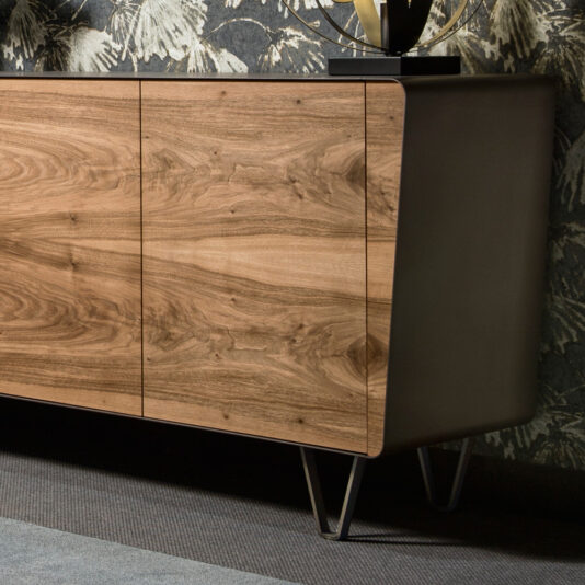 A mid-century style walnut sideboard, distinguished by its modern wooden frame in a dark finish and three vertical panels showcasing a natural wood grain look. This elegant piece is elevated with metal hairpin legs and is set against an eye-catching patterned wall adorned with leafy designs, partially visible in the background.
