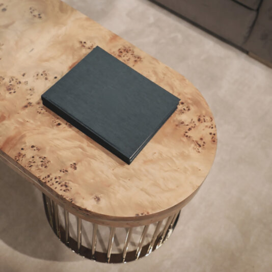 Modern Burl Veneer Coffee Table A sleek, black book is neatly placed on the Modern Burl Veneer Coffee Table with rounded edges and a polished, wood-grain finish. The table stands on a metallic, cylindrical base, resting on a beige carpeted floor next to a plush sofa.
