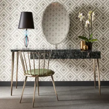 The Modern Dressing Table With Oval Mirror showcases a sleek design with a black marble-top surface and gold legs. It is paired with an elegant gold chair adorned with a green cushion. To complete the chic setup, there is a stylish lamp featuring a colorful glass base and a potted orchid, all beautifully contrasted against geometric-patterned wallpaper.