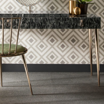 The Modern Dressing Table With Oval Mirror showcases a sleek, black textured surface and slim gold legs, standing against a wall adorned with geometric diamond-patterned wallpaper. Displayed on the table are elegant gold cylindrical objects and a small potted plant. A green velvet chair with matching gold legs is positioned in front of the table.