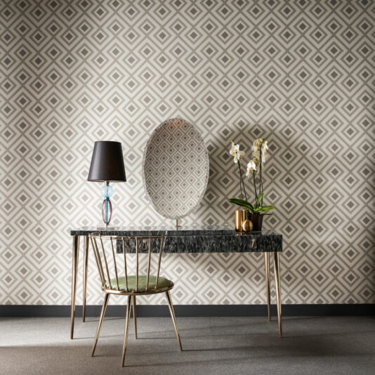 The Modern Dressing Table With Oval Mirror features a sleek black marble top and is paired with a gold chair. The table is adorned with a black lamp that has a transparent base and holds a gold pot containing white orchids, all set against geometric patterned wallpaper in shades of beige and gray.