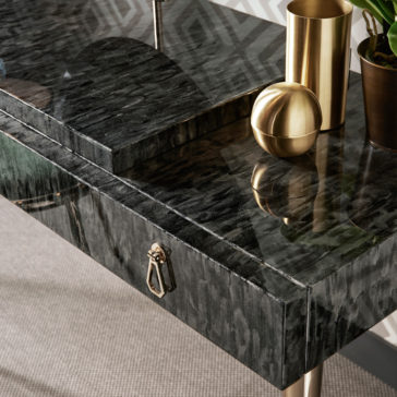 A close-up of the Modern Dressing Table With Oval Mirror reveals its dark, glossy marble surface with a sleek, reflective finish. The table features a small drawer adorned with a metallic handle. On top of the table sits a gold-colored cylindrical container, a spherical decor piece, and a potted plant.