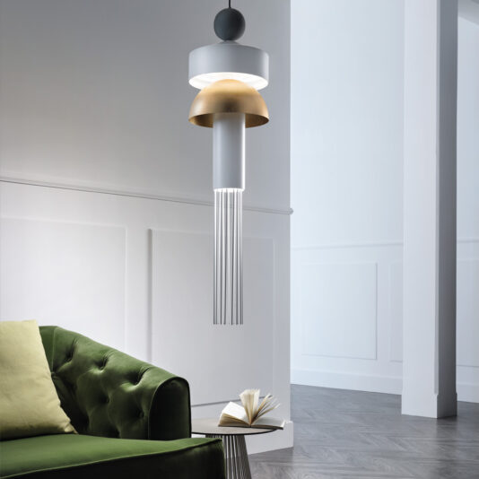 A modern interior features a green velvet sofa with a partially open book placed on the armrest, accompanied by the **Modern LED Pendant Light** showcasing metallic gold and white elements with thin vertical rods. The background boasts white paneling and wood flooring.