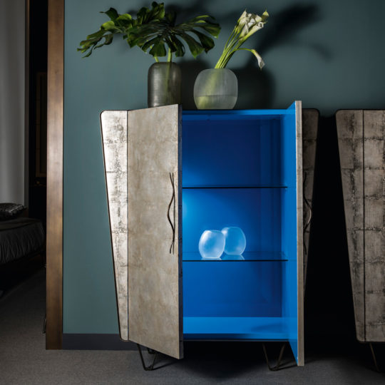 The Modern Silver Leaf Cabinet features metallic textured doors, one of which is open to display a blue interior and glass shelves. Inside the cabinet, two clear glass vases are placed on a shelf. On top of the cabinet, there are two green vases containing leafy plants.