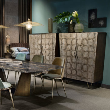 Modern Silver Leaf Cabinet A modern dining room showcases a dark wooden table surrounded by six green upholstered chairs. Behind the table, two Modern Silver Leaf Cabinets stand, adorned with decorative vases on top. The teal walls feature two framed botanical prints, and an overhead pendant light illuminates the space.