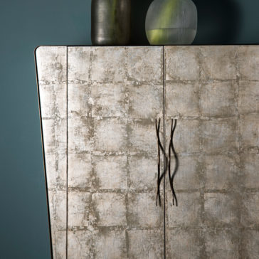 Modern Silver Leaf Cabinet The Modern Silver Leaf Cabinet, boasting textured wooden panels with square patterns and curved metal handles, stands side by side. Two decorative vases of varying shapes and colors are artfully placed on top, set against a plain, dark-colored wall.