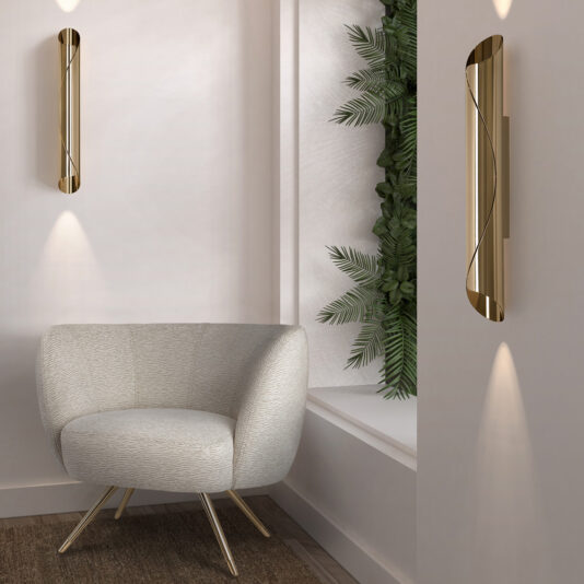 A contemporary, minimalist room showcasing an upholstered beige Modern Tub Armchair with metallic legs, complemented by two sleek vertical gold wall sconces that emit a soft light. Behind the chair, green foliage adorns the wall, bringing a hint of nature into the space.