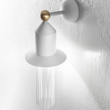 The Modern White LED Wall Light showcases a minimalist design with a white conical shade, topped by a small golden sphere and adorned with thin dangling metallic strands. Mounted on a textured wall, this light casts a soft glow, creating a sleek, contemporary appearance.