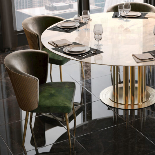 A round, modern dining table with a polished, light-colored surface and a golden base is set for dining with plates, napkins, silverware, and empty glasses. The chairs around the table feature dark green velvet seats and backs from the Quilted Dining Chair collection, supported by gold-colored legs.
