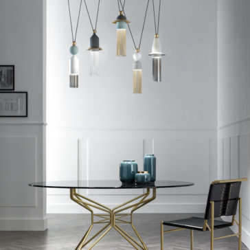 A contemporary dining space features a round black glass table with a gold geometric base, matched with a chic black chair adorned with gold accents. Overhead, the Set Of Modern Pendant Lights is artistically suspended, complemented by blue and white ceramic vases on the table. The walls are white with wainscoting.