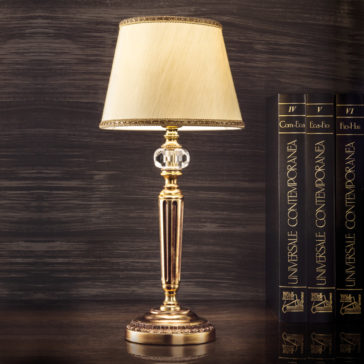 Traditional Gold Plated Table Lamp A **Traditional Gold Plated Table Lamp** with a cream-colored shade and an ornate crystal base sits on a wooden surface. Next to the lamp, there are several large books with black and gold covers, titled