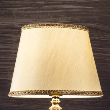 Traditional Gold Plated Table Lamp A close-up of the Traditional Gold Plated Table Lamp reveals its beige shade adorned with gold trim. The lamp’s ornate, gold-colored base enhances its elegance, set against a backdrop of dark textured wood or painted surface.