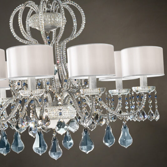 The Traditional Italian Crystal Chandelier is an ornate light fixture featuring white cylindrical lampshades and intricate detailing. It boasts elaborate metalwork adorned with shimmering clear and blue crystal pendants, creating a luxurious appearance against a muted background.