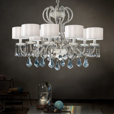 Traditional Italian Crystal Chandelier A Traditional Italian Crystal Chandelier with eight white lampshades and ornate crystal details hangs from the ceiling. The chandelier, adorned with intricate metallic scrollwork and multiple blue crystal teardrops, casts an elegant ambiance over a dimly lit room featuring modern decor.