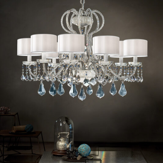 A Traditional Italian Crystal Chandelier with eight white lampshades and ornate crystal details hangs from the ceiling. The chandelier, adorned with intricate metallic scrollwork and multiple blue crystal teardrops, casts an elegant ambiance over a dimly lit room featuring modern decor.