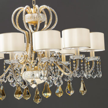 The Classic Crystal Chandelier With Shades is a luxurious piece featuring an elegant gold and crystal design with eight arms adorned with cream-colored lampshades. The chandelier is decorated with sparkling crystal beads and large teardrop-shaped pendants, creating a sophisticated ambiance against a dark gray background.