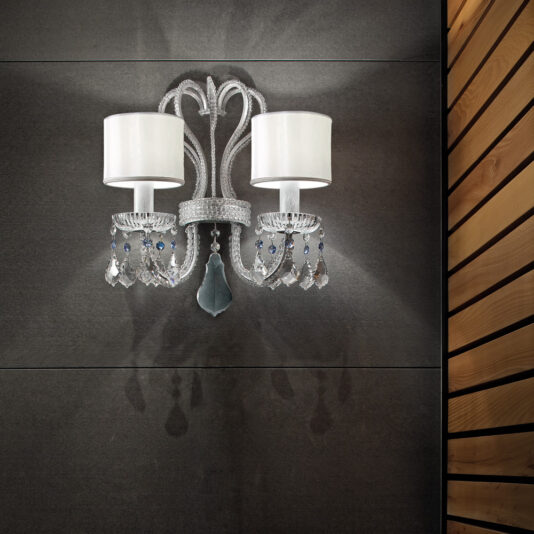 The **Traditional Double Crystal Wall Light**, featuring two white lampshades adorned with crystals and a teardrop pendant, is mounted on a dark gray wall. To the right, a wooden wall showcases a vertical slat design. This elegant double crystal wall light emits a soft glow, casting delicate shadows on the surface.
