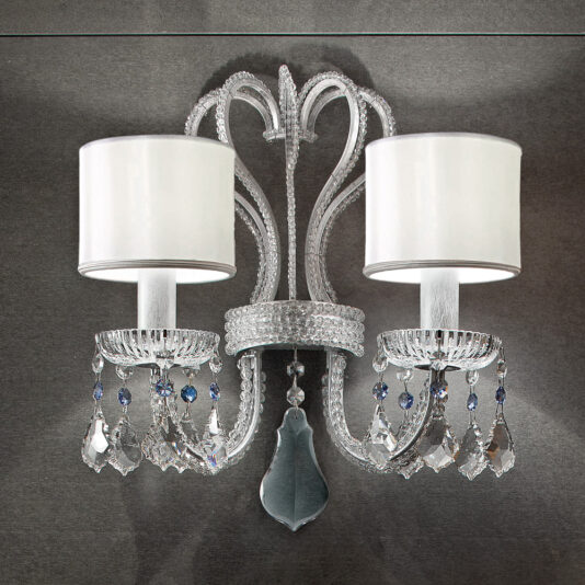 The Traditional Double Crystal Wall Light features a twin-arm design with a silver finish and is adorned with two white cylindrical lampshades. This sconce showcases ornate detailing and dangling crystal accents, all set against a dark gray background.