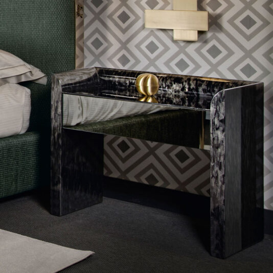 A sleek, reflective black marble console table with modern lines stands in a contemporary room. Positioned against a gray and white geometric-patterned wallpaper, it's complemented by a round, golden decorative object. Nearby, the exquisite Black Mother Of Pearl Bedside Cabinet accentuates the partially visible bed.