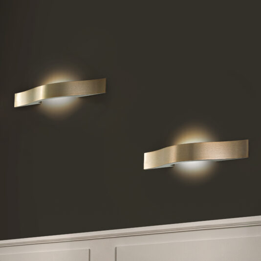 Two Contemporary Burnished Brass Wall Lights with wave shapes softly illuminate a dark wall. The warm glow enhances the ambiance above the white wainscoting at the bottom of the image.