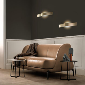 A modern living room showcases a tan leather sofa with a dark throw. Contemporary Burnished Brass Wall Light fixtures illuminate the upper wall, while white paneling adorns the lower section. Black side tables hold decor and books on the light wood floor below.