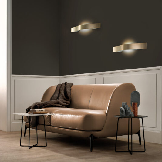 A modern living room showcases a tan leather sofa with a dark throw. Contemporary Burnished Brass Wall Light fixtures illuminate the upper wall, while white paneling adorns the lower section. Black side tables hold decor and books on the light wood floor below.