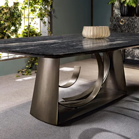 A Large Modern Bronze Dining Table with a dark marble top and unique curved base sits elegantly on a gray carpet. Sunlight through a nearby window with green foliage illuminates the room, casting a glow on the decorative bowl atop the table.
