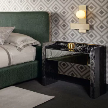 Modern bedroom with geometric wallpaper featuring a **Black Mother Of Pearl Bedside Cabinet** and a brass light fixture above it. The bed has a green upholstered headboard and grey bedding with decorative pillows. The floor is carpeted in a dark shade, and a grey rug is partially visible.