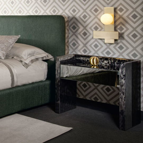 Modern bedroom with geometric wallpaper featuring a **Black Mother Of Pearl Bedside Cabinet** and a brass light fixture above it. The bed has a green upholstered headboard and grey bedding with decorative pillows. The floor is carpeted in a dark shade, and a grey rug is partially visible.