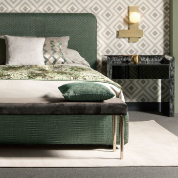 A modern bedroom highlights a green upholstered bed adorned with patterned pillows and a leafy bedspread. At the foot of the bed is a sleek bench topped with a green cushion. The decor is completed with geometric wallpaper, brass wall sconce, and the Black Mother Of Pearl Bedside Cabinet.