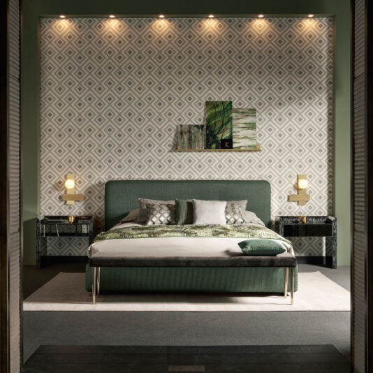 A contemporary bedroom boasting a spacious bed complemented by numerous pillows, a green upholstered headboard, and an elegant bench at the foot. Geometric wallpaper adds texture, flanked by two Black Mother Of Pearl Bedside Cabinets topped with lamps, while a wall painting elevates the aesthetic. Warm ceiling lights softly illuminate the room.