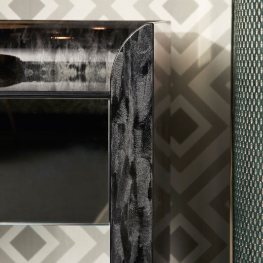 Close-up of the Black Mother Of Pearl Bedside Cabinet, showcasing its glossy, textured black finish. The backdrop features a geometric wallpaper pattern in shades of gray and white, accented by a textured dark green panel on the right edge.