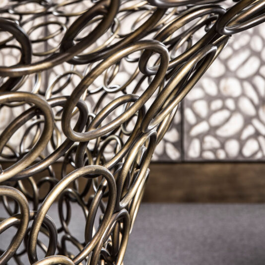 A close-up photo captures the intricate design of the Bronze Rings Round Glass Dining Table, featuring abstract bronze rings that form a complex, woven pattern. These interlocking metal rings elegantly contrast against a textured wall with an irregular stone-like pattern, enhancing the modern aesthetic of the setting.