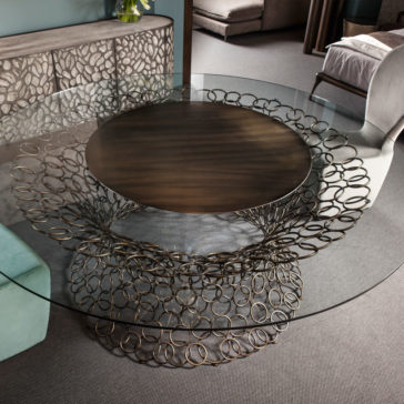Bronze Rings Round Glass Dining Table The Bronze Rings Round Glass Dining Table, featuring a distinctive base composed of interlinked bronze rings, is the star of the room. A large dark wooden centerpiece adorned the table, complemented by modern decor including a patterned sideboard, upholstered chairs, and plush carpeting.