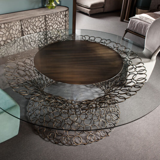 The Bronze Rings Round Glass Dining Table, featuring a distinctive base composed of interlinked bronze rings, is the star of the room. A large dark wooden centerpiece adorned the table, complemented by modern decor including a patterned sideboard, upholstered chairs, and plush carpeting.